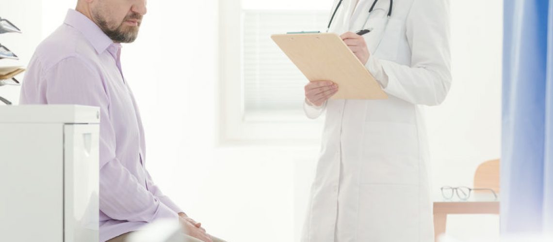 Sick man sitting next to a urologist in white uniform with stethoscope while making a diagnosis